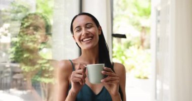 Genç ve güzel bir kadın kafasında fincanı tutuyor, taze demlenmiş sabah çayının ya da kahve içkisinin tadını çıkarıyor, dinlenmek ve tadını çıkarmak için zaman ayırıyor, yaz günü pencerenin dışındaki bulanık yeşilliğin tadını çıkarıyor.