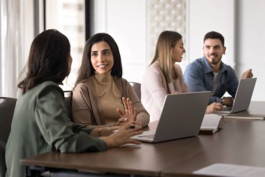 Pozitif, genç, güzel, Latin, profesyonel bir kadın iş yerinde iş arkadaşıyla konuşuyor. İspanyol iş arkadaşları çalışma masasında toplanıyor, proje görevlerini, takım çalışmasını, işbirliği stratejisini tartışıyorlar.