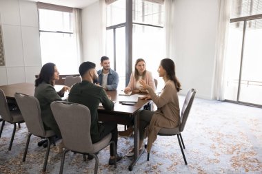 Takım toplantısında konuşan kendinden emin genç Latin bayan iş adamı ofis işbirliği çalışma alanında büyük bir masada oturuyor, iş arkadaşlarıyla konuşuyor, takım çalışmasını tartışıyor, grup işbirliği yapıyor.