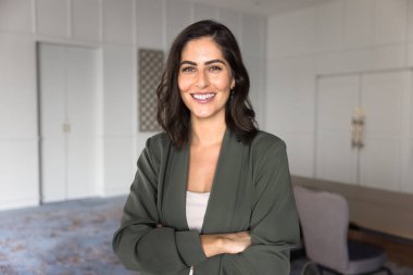 Kendine güvenen güzel Latin yönetici kadın elleri çaprazlanmış kameraya bakıyor, toplantı odasında tek başına duruyor. Mutlu genç İspanyol iş kadını portresi