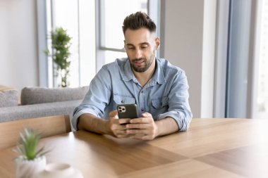 Ciddi odaklı İspanyol genç adam ofis masasında cep telefonuyla yazı yazıyor, akıllı telefonda e-iş uygulaması kullanıyor, internette sohbet ediyor, internette geziniyor, sosyal medya, sipariş veriyor, alışveriş yapıyor.