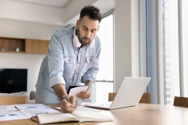 Başlangıç için araştırma yapan ciddi odaklı proje yöneticisi, kağıt belgeleri, pazarlama raporları, dizüstü bilgisayarla masaya notlar yazmak, finansal verileri analiz etmek, istatistiksel bilgiler