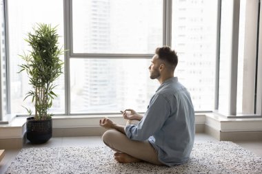 Huzurlu odaklı genç Latin yogi adam evde meditasyon yapıyor, zen ellerini ve meditasyon pozunu tutuyor, geniş pencereli, şehir manzaralı, kapalı gözlerle oturuyor, mantraları dinliyor, konsantre olmaya çalışıyor.