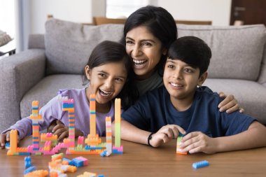 Neşeli Hintli kardeş çocuklar ve genç anne inşaat oyununda aile portresi için poz veriyorlar, renkli oyuncak kulelere sarılıyorlar, bina blokları inşa ediyorlar, uzağa bakıyorlar, gülümsüyorlar, gülüyorlar.