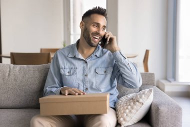 Elinde karton paketler olan yakışıklı Latin tüketici, evde cep telefonuyla konuşuyor, gülümsüyor, gülüyor, cep telefonuyla internet e-ticaret mağazasından alışveriş yapmayı tartışıyor, teslimat siparişi veriyor.