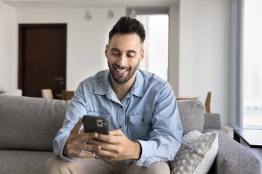 Mutlu yakışıklı İspanyol genç adam internet teknolojisini cep telefonunda kullanıyor, cep telefonunda yazıyor, internette sohbet ediyor, kanepede oturuyor, sosyal medyada cihazlarda geziniyor, gülümsüyor, gülüyor.