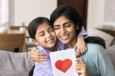 Mutlu Hintli anne kucaklayan kız çocuğu, kırmızı kalpli el yapımı tebrik kartı tutuyor, anneler gününü kutluyor, 8 Mart bayramı, kapalı gözlü çocuğu kucaklıyor.