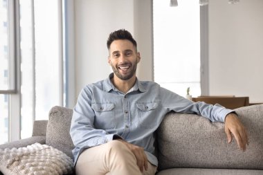 Mutlu, çekici, genç İspanyol ev sahibi adam oturma odasında koltukta oturuyor, kameraya bakıyor, gülümsüyor, gülüyor, neşe ifade ediyor, ev portresi için poz veriyor, rahat koltukta dinleniyor.