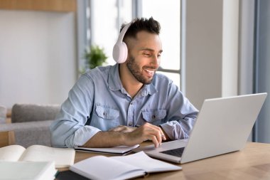 Kulaklıklı mutlu yetişkin Latin öğrenci laptopta ders çalışıyor, masada kitaplar açıyor, konferans seyrediyor, konferans sohbetleri yapıyor, eğitim için teknolojiyi kullanıyor.