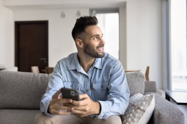 Rüya gibi yakışıklı Latin adam evde online iletişim için alet kullanıyor, akıllı telefon tutuyor, pencereye bakıyor, gülümsüyor, iş için internet teknolojisini düşünüyor.