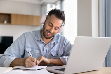 Olumlu yetişkin İspanyol öğrenci kulaklık takıyor, çevrimiçi konferans izliyor, web sitelerini eğitiyor, not alıyor, kitap yazıyor, açık kitaplarla evde oturuyor.