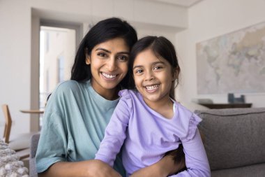 Mutlu, genç, Hintli bir anne kucağında tatlı kızı, koltukta oturuyor, kameraya bakıyor, gülümsüyor, çocuğa sevgiyle sarılıyor, şefkatle, şefkatle, anneliği destekliyor.