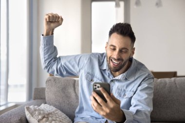 Mutlu, aşırı mutlu, yakışıklı İspanyol adam evde cep telefonu kullanıyor, kazanan el kaldırıyor, kazanan el hareketi, kazanan ödül, çevrimiçi başvuruda harika haberler alıyor.