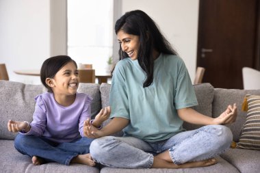 Neşeli Hintli anne ve şirin kız evde yoga yapıyor, meditasyon ve zen eller için poz veriyor, rahat koltukta oturuyor, konuşuyor, sohbet ediyor, gülüyor, eğleniyor.