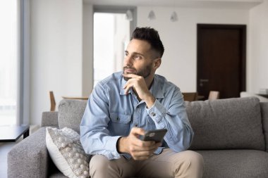 Düşünceli, rüya gibi, çekici İspanyol bir adam cep telefonu tutuyor ve başka tarafa bakıyor, rahat bir kanepeye oturuyor, çeneye dokunuyor, sosyal medyayı karıştırıyor, internet iletişimini düşünüyor.