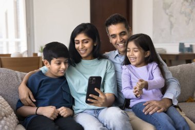 Mutlu Hintli anne ve baba akıllı telefon kullanıyor, küçük sevimli kardeş çocuklara kanepede sarılıyorlar. Anne çevrimiçi içerik gösteriyor, öğrenme uygulaması, çocuklara ve babalara oyun