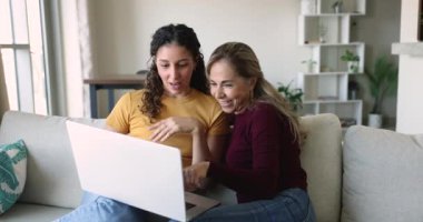 Mutlu büyük anne ve genç yetişkin kız evde dizüstü bilgisayar kullanıyorlar, monitörü gösteriyorlar, konuşuyorlar, gülümsüyorlar, online alışveriş, satın alma, internet iletişimini tartışıyorlar.