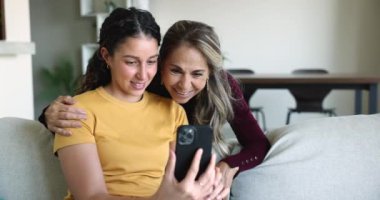 Olumlu genç Latin kız ve olgun anne cep telefonundan online video içeriği izliyorlar, neşeli oluyorlar, gülüyorlar, internet iletişiminden keyif alıyorlar, evde teknoloji var.