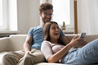 Neşeli, çekici, genç eşler rahat koltukta dinleniyor, cep telefonu videoları kullanıyor, akıllı telefonda komik videolar izliyor, hafta sonunu evde geçiriyor, yeni mobil uygulamalarla eğleniyor, internet eğlencesinin tadını çıkarıyor.
