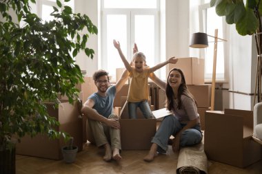 Küçük kız karton kutudan atlıyor, eğleniyor, taşınma gününün tadını çıkarıyor, gülümsüyor genç ebeveynlerle kameraya bakıyor, karton kutuların yanında oturuyor. Banka kredisi, yeni ev, yeni ev.