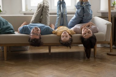 Aile kanepede baş aşağı yatıp gülüyor ve kameraya bakıyor. Kaygısız, aktif, küçük kızı olan, eğlenen, rahat koltukta bacaklarıyla egzersiz yapan ebeveynler. Boş zaman