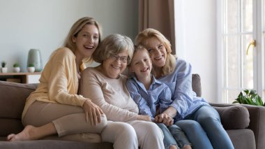 Farklı yaşlardaki neşeli kadın akrabalar, nesiller boyu ev portresi. Mutlu küçük kız, genç anne, olgun büyükanne, yaşlı büyük anne kanepeye yakın oturuyor, sarılıyorlar.