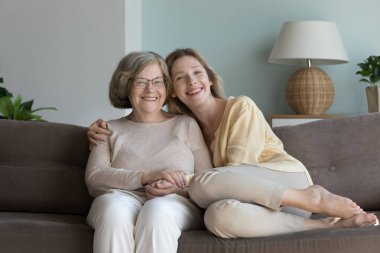 Neşeli yaşlı bir büyükanne ve güzel bir torun kız el ele tutuşuyor, sarılıyor rahat bir kanepeye oturuyor, kameraya bakıyor, gülümsüyor, gülüyor. Aile fotoğrafı.