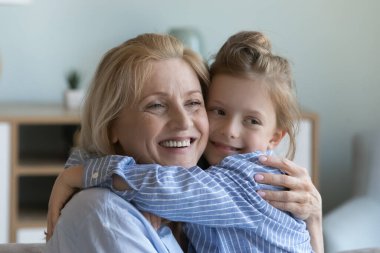 Neşeli bir büyükanne torun kızına sevgi, ilgi, şefkat, şefkat ile sarılıyor, dişlek bir gülümsemeyle başka tarafa bakıyor, yanak yüzlü bir çocuğu kollarına alıyor, dokunuyor.