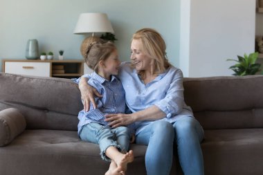 Orta yaşlı mutlu bir büyükanne tatlı bir torunla konuşuyor, evde bir kızla kanepede dinleniyor, çocukla sohbet ediyor, gülümsüyor, gülüyor, güvenin tadını çıkarıyor, arkadaşlığın tadını çıkarıyor.