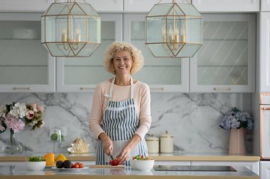 Mutlu orta yaşlı aşçı blogcu kadın apronlu modern mutfak içlerinde taze sebzeler kesiyor, masada duruyor, kameraya bakıyor, gülümsüyor. Sağlıklı beslenme. İçerideki portre