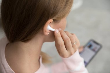 Genç beyaz alet kullanıcısı kadın kulaklığa dokunuyor, arka planda akıllı telefonu tutuyor, çevrimiçi medya içeriğini izliyor, video konuşmaları yapıyor. İnternet iletişimi