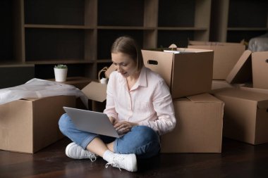 Taşınmaya dizüstü bilgisayar kullanan, boş bir dairedeki kutuların üstündeki bilgisayarı kullanan, taşınma, paketleme ve ulaşım hizmetleri için internet arayan pozitif kiracı kadın.
