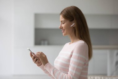 Oturma odasında duran çekici genç bir kadın akıllı telefon kulaklığı takıyor. Akıllı telefon ekranına bakıyor. Gülümsüyor, favori müziğin tadını çıkarıyor, şarkı seçiyor, podcast 'i dinliyor, modern teknoloji kullanıyor.