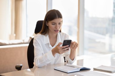 Ciddi meşgul, uzaktan kumandalı bir kadın internet iletişimi için akıllı telefon kullanıyor, iş yerindeki defterin başında oturuyor, ofisten çalışıyor, internette sohbet ediyor.,