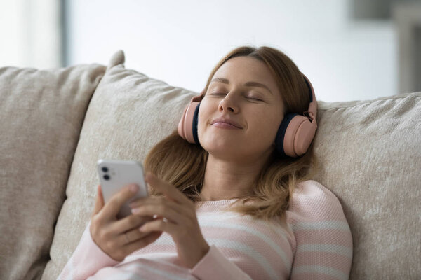 Расслабленный беззаботный тысячелетний гаджет пользователь женщина носит большие наушники, отдыхает на домашнем диване с закрытыми глазами, с помощью онлайн-приложения, плеер на мобильный телефон, слушая успокаивающую музыку из Интернета