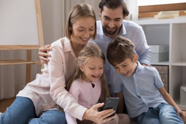 Mutlu bir çift ebeveyn ve iki küçük çocuk cep telefonlarıyla video görüşmesi yapıyorlar, sohbet ediyorlar, aile selfie 'si çekiyorlar, evde dinleniyorlar, ekrana bakıyorlar, webcam izliyorlar, gülüyorlar.