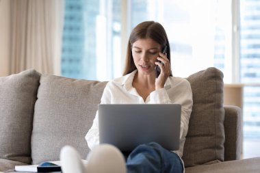 Odaklanmış genç laptop kullanıcısı serbest çalışan kız cep telefonuyla konuşuyor, dijital cihazlarla evdeki koltuklarda dinleniyor, iş için internet iletişimi için kablosuz teknoloji cihazları kullanıyor.