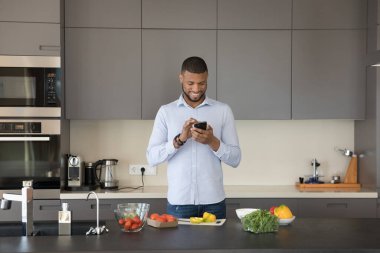 Neşeli yakışıklı Afrikalı genç adam internette akıllı telefondan yemek tarifini tarıyor, yemek bloguna danışmanlık yapıyor, resim çekiyor, salata pişiriyor, sebze kesiyor, internetten sipariş veriyor.