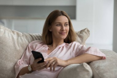 Güzel bir kadın elinde akıllı telefonuyla koltukta oturup uzaktan bakıyor evde yalnız başına dalgın dalgın bakıyor, sosyal medya ağını, perakende e-hizmetlerini, elektronik alışverişi, mobil uygulama kullanıcısını kullanıyor.