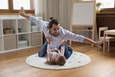 Mutlu, genç bir baba ve küçük bir kız çocuk odasında eğleniyor. Gülen kız sıcak zeminde dinleniyor, sırt üstü uzanıyor, babasının çocuk üzerinde uçmasını destekliyor.