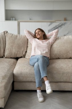 Mutlu kadın, modern evdeki rahat oturma odasındaki rahat koltukta dinlenmenin keyfini çıkarıyor. Yumuşak yastıklara yaslanmış gülümsüyor, ellerini başının arkasına koyup, evde kaygısızca vakit geçiriyor.