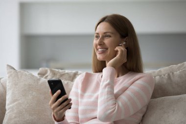 Neşeli rüya gibi bin yıllık akıllı telefon kullanıcısı kadın kulaklığa dokunuyor, cep telefonunu tutuyor, telefon görüşmesi yapıyor, online medya uygulaması ile müzik dinliyor, ev koltuğunda dinleniyor.