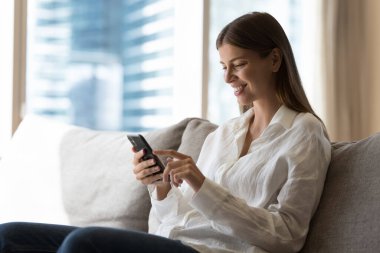 Akıllı telefondan mesaj yazan, evdeki koltukta oturan, internetten alışveriş yapan, internetten iletişim kurmak için alet kullanan, ekranda gülümseyen güzel, akıllı telefon kullanıcısı kadın.