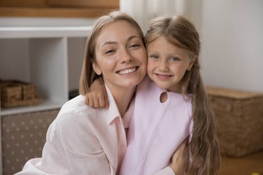 Mutlu genç anne ve güzel küçük kız yanaklarına dokunarak sarılıyor, kameraya bakıyor, gülümsüyor, sevgiyi ifade ediyor, şefkati ve memnuniyeti ifade ediyor. Anne ve sevimli çocuk evde portre resmi çekiyorlar.