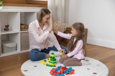 Mutlu genç anne ve küçük kız el çırpıyor, ahşap yapı taşları üzerinde beşlik çakıyor, ısıtmalı zeminde oyuncak bina yapıyor. Bakıcı ve çocuk evde oyun oynuyorlar.