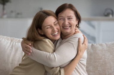 Neşeli, olgun bir anne. Kucaklayan, evdeki koltukta kızına sarılan, gülen, yetişkin çocuğa sevgiyle sarılan, şefkatle, mutlulukla, gülümseyerek, aile bağlarından keyif alan.