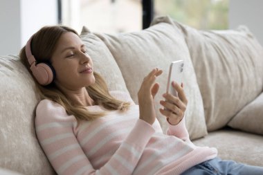 Rahatlamış, milenyum akıllı telefon kullanıcısı büyük kablosuz kulaklıklı kadın müzik dinliyor, şarkılar dinliyor, cep telefonundan radyo dinliyor, video görüşmesi yapıyor, içeriği izliyor, rahat ev koltuğunda dinleniyor.