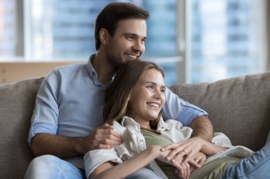 Neşeli, genç bir çift, sevgiyle kucaklaşıyorlar, evde dinleniyorlar, kanepede dinleniyorlar, başka yere bakıyorlar, düşünüyorlar, hayal ediyorlar, planlıyorlar, gülüyorlar, eğleniyorlar, eğleniyorlar, sağlıklı bir ilişki yaşıyorlar.