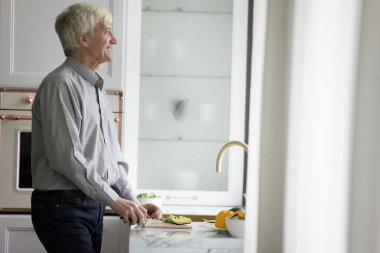Neşeli, rüya gibi kıdemli adam modern ev mutfağında yemek yapmaktan zevk alıyor, taze sebzeleri kesiyor, mutlu bir gülümsemeyle pencereye bakıyor, düşünüyor, hayal ediyor, yemek planlıyor. Sağlıklı beslenme, emeklilik