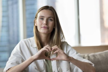 Güzel, genç bir kız romantik bir el kalbi gösteriyor, aşk, romantizm, destek, hayırseverlik jestleri yapıyor, kameraya bakıyor, içeride poz veriyor, olumlu duygular gösteriyor.